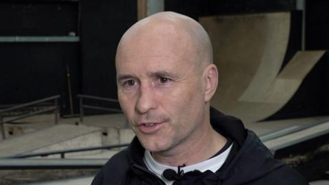 A bald man in a skateboard park wearing a black hoodie.
