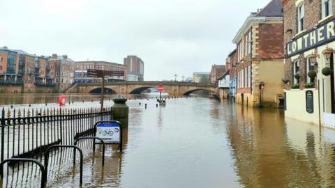 Picture of the River Ouse in York flooded over the banks onto the pavement and road with floodwater rising up the side of the buildings.