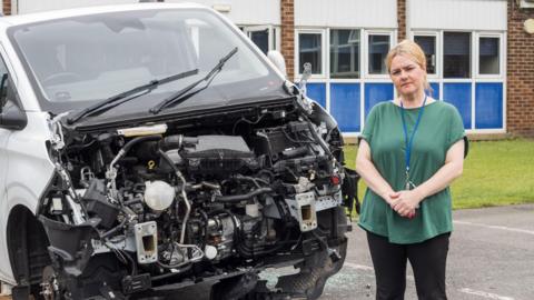 Corrina McEwan, wearing a grey t-shirt, black pants and a work lanyard, stands next to a a van with the bonnet missing at the front, along with its tyres.