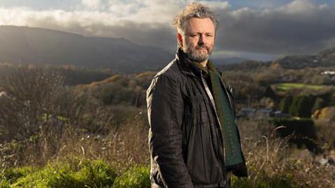 Standing on high ground above a valley, Michael Sheen looks towards the camera with rolling hills and patchwork fields stretching into the distance. He is wearing a brown jacket with a green patterned jumper. Sheen has grey hair and facial hair. Strong afternoon sunlight and heavy clouds create dramatic contrasts across the rural landscape.