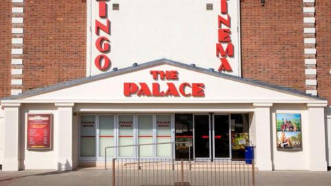A close view of the entrance into the Palace Cinema in Felixstowe. The building is large and made of red bricks. It has a white entrance with numerous doors. Movie posters can be seen either side of the entrance,