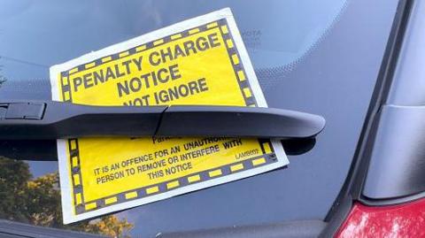 A penalty charge notice on a car, placed under the windscreen wiper. The red paint of the car is visible and there are trees reflected in the windscreen.