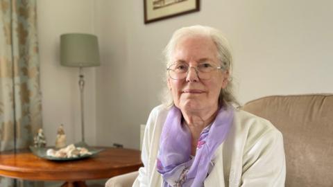 A woman with shoulder-length grey hair wearing a white cardigan and lilac scarf sits in her living room. She is on a light brown armchair and behind her is a brown coffee table with shells on a plate and two statues next to it on the table. There is a lamp with a light-green shade in the corner of the room, next to light gold and green floral patterned curtains. 