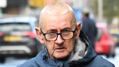 William Ball. He has very short white hair, wearing black glasses and a blue hooded raincoat.