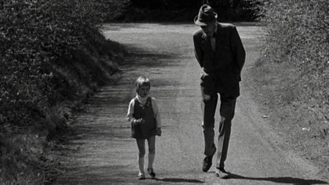 Black and white image of a young girl walking alongside a tall man in a hat