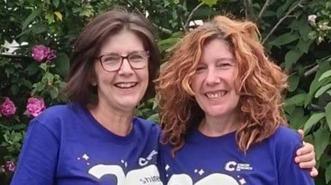 Jill and Jan smile as they embrace with their arms around each other for the camera. They both wear blue Cancer Relief UK tops as they stand in a garden with pink roses and green shrubs behind them. Jill has dark shoulder-length hair and wears glasses. Jan has wavy ginger hair.