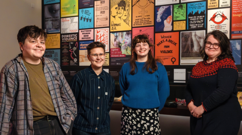 Four people standing in front of an exhibition of feminist posters. The first person has short brown hair and is wearing a green T-shirt and plaid shirt. The next person has short brown hair and glasses and is wearing a navy striped shirt. The next person has long brown hair and is wearing a blue jumper and a floral skirt and the last person has long brown hair and is wearing a black and red jumper-dress.