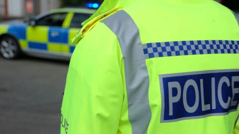 Back of a police officer in a yellow hi-vis jacket with POLICE written in grey. The jacket also has a line of blue and grey checks on it. There is a blue and yellow checked police car in front of the officer.