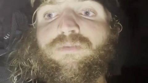 Lachlan has grown a big curly blond beard. It is a close up of his face and darkness surrounds him.