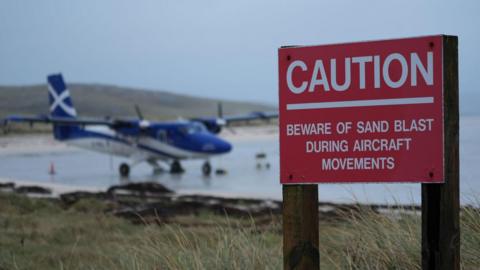 A red warning sign at Barra's beach airport. There is a plane in the background.