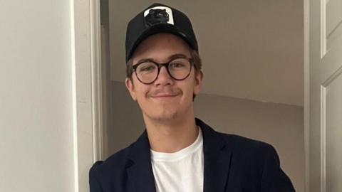A young man, Freddie Penrose, is wearing a cap and glasses leans against a door frame. He is wearing a suit jacket.
