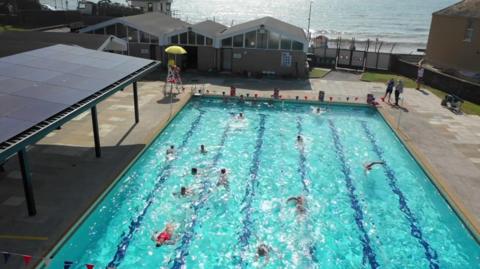People swimming in a swimming pool. There is a lifeguard sat on a tall chair with a yellow umbrella. In the background is the sea. 