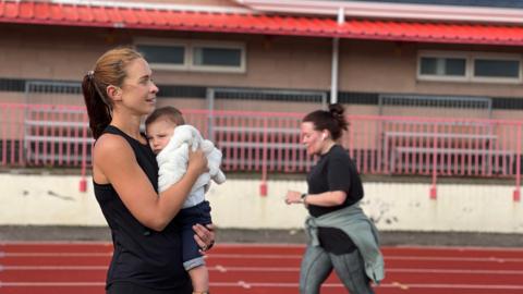 The coach holds a baby as she looks to right. Behind her is a running track and a mum is running behind her to her right. The coach is in black exercise kit and her hair is in a ponytail. The baby is wearing a white coat and blue shorts. The mum running has her hair in a bun and is wearing a black exercise top with grey joggers and has a grey coat tied around her waist.