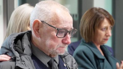 Geoff Burrow in a grey shirt and tie with a blue jumper, grey coat and dark glasses has short grey hair and a goatee.