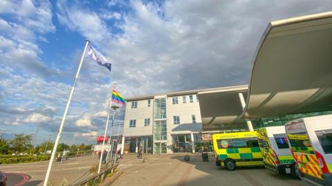 Darent Valley Hospital in Dartford, England, with people visible in the background and the NHS and Gay Pride flags outside.