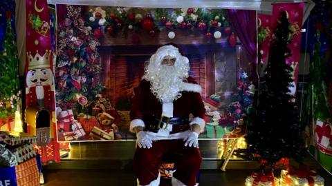 A Santa surrounded by Christmas presents and a decorative Christmas backdrop.