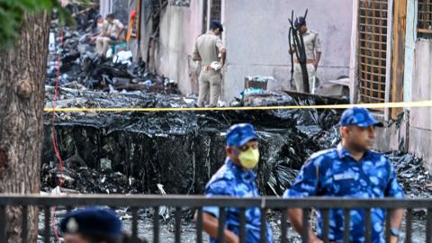 Officials in masks look on while standing outside yellow tape cordoning off the burnt-out crash site, with other officials at the wreckage inspecting, on 13 June