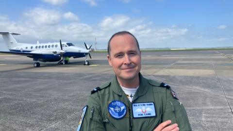 Air Cdre Rob Caine wearing a green pilots jumpsuit with blue badges. He has very short dark hair and is clean shaven. Behind him on the runway is a King Air Avenger training aircraft. It is white and blue with two large propellers on the wings. 