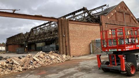 Bricks pile up at an entrance of of a former bus depot. The yard can be seen. Old wall structures of former buildings surround the yard area. 