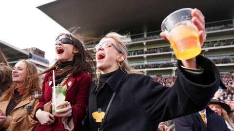 Three young women cheer with their mouths wide open as they watch the conclusion of a race on Gold Cup day at the Cheltenham Festival. A packed grandstand is visible behind them