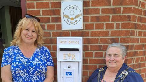 Sharon Sutcliffe has blond hair and sunglasses on top of her head. She is wearing a blue dress with a flower pattern and is standing on the left-hand side  of a white postbox, which is mounted on a brick wall with a sign above it. To the right stands Hull's Lord Mayor Councillor Cheryl Payne who has short grey hair and dark blue top with the silver mayoral chains around her neck.