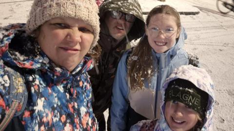 A selfie picture of a woman in a beige woolly hat and a red black and white patterned jacket. She is scrunching up her nose and mouth to the camera, as if in disgust. Crouched behind her is a man in glasses wearing a green jacket with the hood up, a smiling girl with glasses, long blonde hair and a blue jacket, and a shorter girl with a black hat and a light-coloured hood up. They are standing in a snowy street.