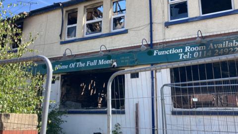 A damaged building is shown behind temporary metal safety fencing. Several upper-floor windows are broken, and the roof structure appears heavily damaged. A green sign on the building reads “Home Of The Full Monty” and includes text advertising a function room with a telephone number. Vegetation is visible along one side of the building.