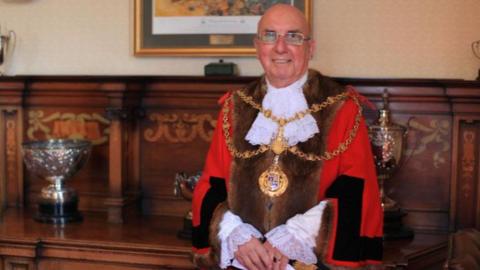 A man wearing mayoral robes and a medal, and glasses, stood in front of a wooden panelled wall with trophies on a wooden surface.