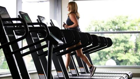 Generic photo of a woman in a gym using a running machine in a row of the machines. There are large glass windows with views of trees