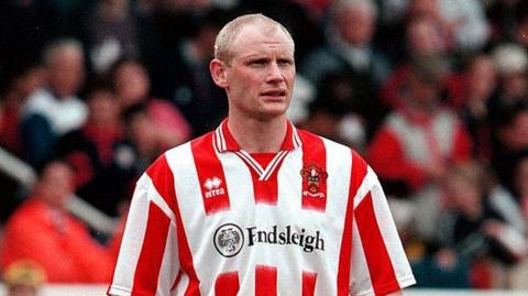 Neil Grayson standing on the pitch wearing a Cheltenham Town shirt in 2000