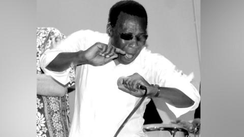 Ray King in stage in an black-and-white image. He is wearing a white shirt and had dark glasses on. He is holding a microphone in his left hand.