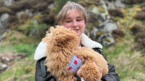 A woman with tied-back hair, wearing a brown leather jacket, is holding a light brown coloured dog in her arms.