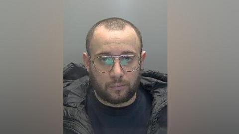 Andi Aliaj is looking at the camera in a police mug shot. He has very short, cropped hair and a brown beard and moustache. He is wearing spectacle and a blue pullover and black padded jacket
