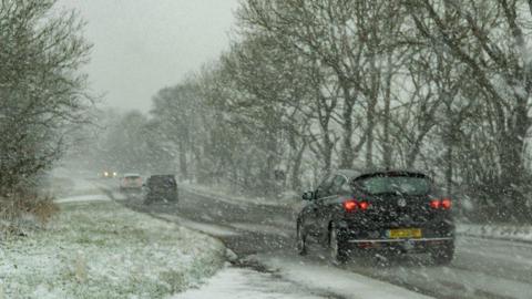 Several cars drive on a road during snow. Snow can be seen falling and settling on the ground and we can see the cars have their lights on.