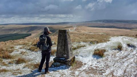 A person standing next to a trig point on a bare moor lightly dusted in snow