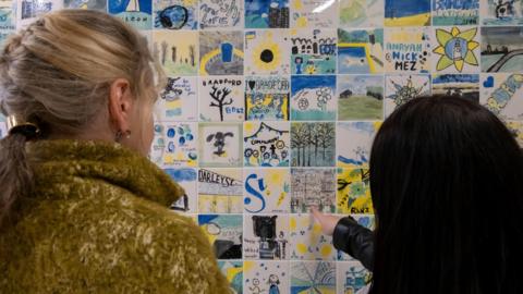 Two people are looking at a wall covered in small, colourful tiles with drawings and text.