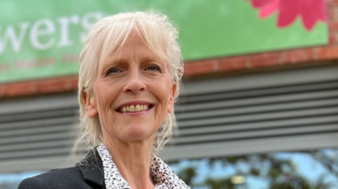 Kate Thomas stands outside her store, Funky Flowers, she is looking at the camera and smiling, her long white hair is tied back in a plait and she has a fringe framing her face. Kate wears a black blazer, with a whit eand black polka dot blouse and grey waistcoat with wooden buttons done up, underneath. Her store is slightly blurred behind her, the Funky Flowers sign is above the doorway, it is a green banner, with the word Funky in pink and Flowers in white, there are plants lining the entryway to the shop.