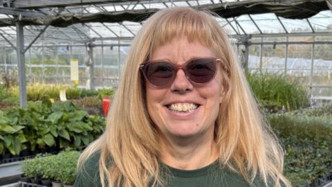 A smiling Heather Lewis standing in the plant nursery with tables full of plants in the background. She has long blond hair and dark glasses.