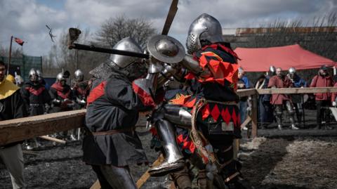 Two people wearing medieval suits of armour engaged in combat, with a crowd of people watching behind