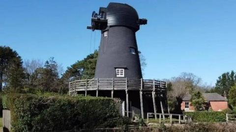 Bursledon Windmill, pictured near greenery and with a house in the background. It has wooden fencing around it and the windmill is painted black.