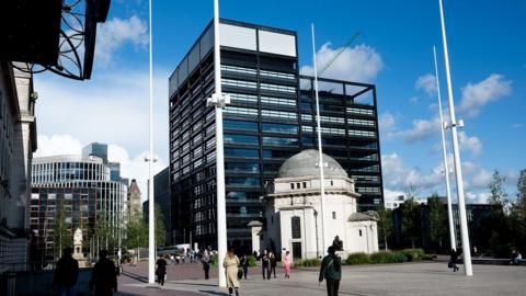 A large glass office building that is about 14 storeys tall. Its exterior is black and white, and there is a roof garden on its back right hand side. In front of it, is a smaller, white domed building.
