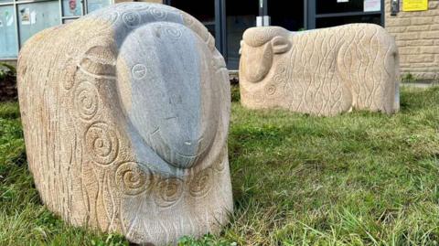 Two large carved stone sheep stand on a patch of grass in front of a brick building, each decorated with swirling patterns cut into their bodies. One sheep is darker and more detailed, while the other is lighter and simpler, with signs and windows from nearby offices visible behind them.