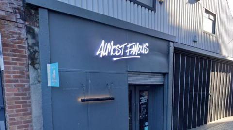 A neon sign for 'Almost Famous' on the front of a grey building above the entrance to the restaurant on Parr Street in Liverpool. 