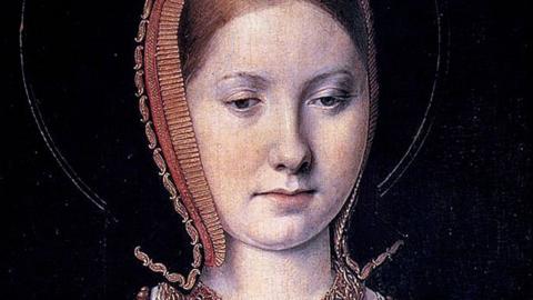 A close-up of a portrait of Katharine of Aragon as a young woman. She is wearing a black headdress edged with red and gold braid and embroidery. Her red hair can be seen pulled back at her forehead. She is lowering her eyes to look down.