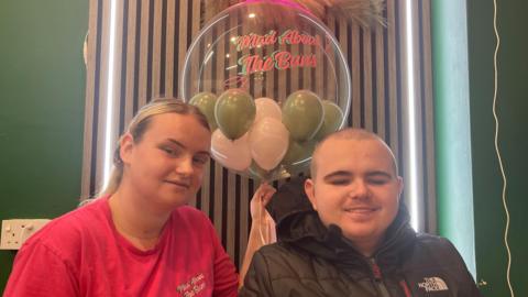 The image shows a blonde woman in a red t-shirt on the left, sitting next to a man in a black coat. The woman is holding a small bunch of green and white balloons below a sign that reads 'Mad about the Buns'.