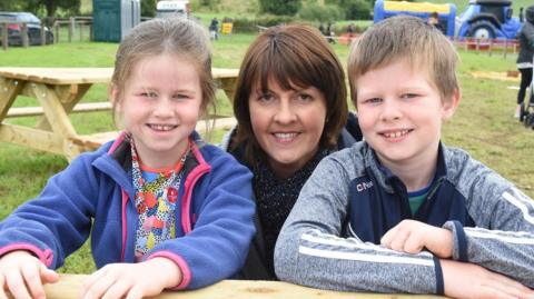 A photo taken a few years ago of Sara Rutledge and her older brother James with their childminder Jenny Bowles. All three are sitting at a picnic bench in a park or rural setting.  A blue tractor and other parked vehicles are in the background