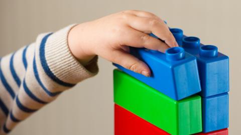 A close-up of a child playing with some colourful building blocks. Their hand and arm is visible, and they are wearing a stripy jumper. You cannot see their face.