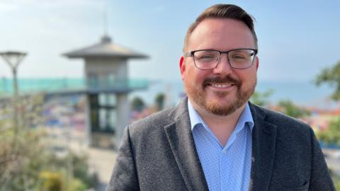 A man with glasses and a short back and sides hair cut and trimmed beard smiling at the camera. He is in his thirties wearing a blue shirt and grey blazer. it is a sunny day and he is at a seafront which is out of focus in the picture.