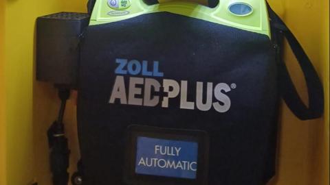 A defibrillator, which is navy with a bright green top, and has a sign that reads "Zoll AED plus fully automatic" on it, sits in a yellow case