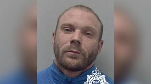A police mugshot of Aaron Moss. He is wearing a blue top and has a beard.